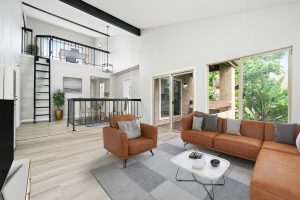 staged living room with couch, chair and coffee table. Wall of windows showing lush landscaping outside