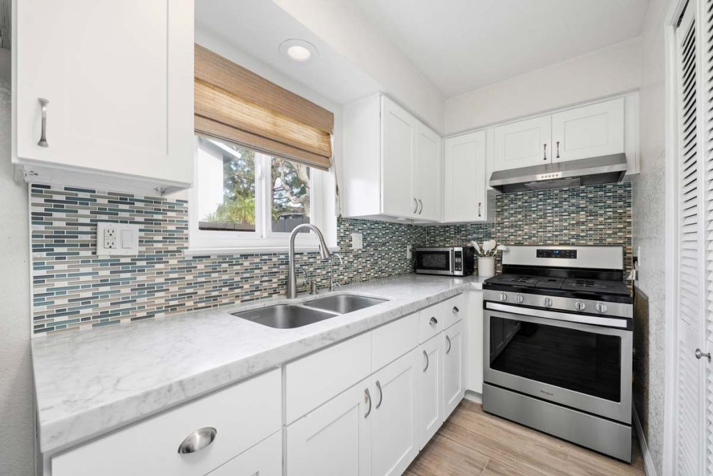 kitchen with white cabinets, light stone counters, multicolored blue and white tiled backsplash, stainless appliances.