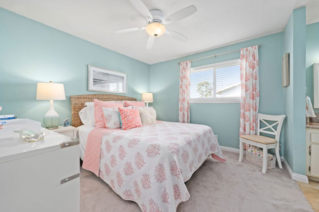 master bedroom with beachy theme, showing light blue walls, white and coral bedspread, light colored carpet and a white ceiling fan.