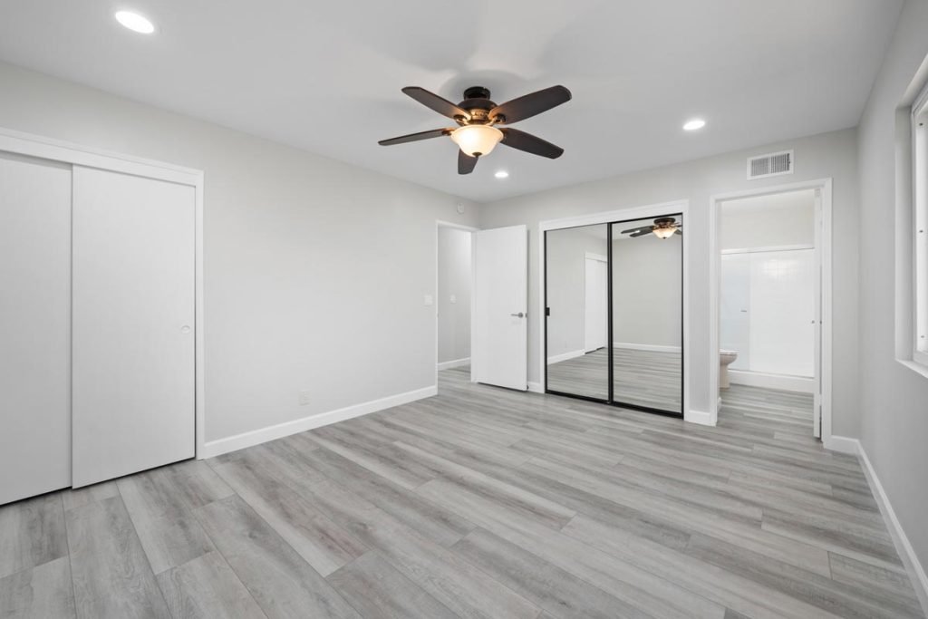 empty master bedroom with gray plank floors, mirrored closet, ensuite bathroom and ceiling fan