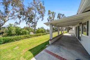 view of grassy area off the back of home for sale, showing a covered patio and large trees in the background.