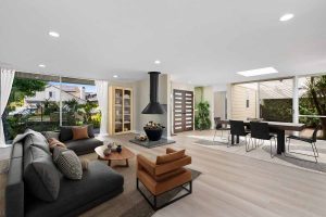 staged living room showing neutral hard wood-looking floors with furniture, a wood burning stove, lots of windows and a mid-century modern style front door.