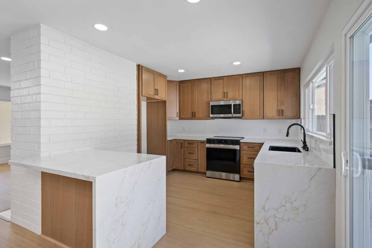 An updated kitchen with light wooden cabinets and white quartz countertops that waterfall over the edge of the counter. Black and stainless fixtures and appliances.