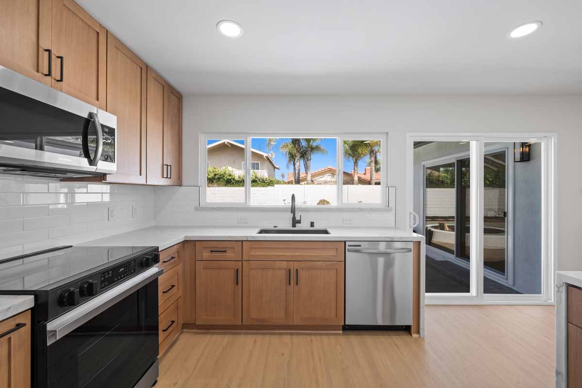 An updated kitchen with light wood cabinetry, white counter tops, and stainless appliances, all looking out through a window to the backyard.