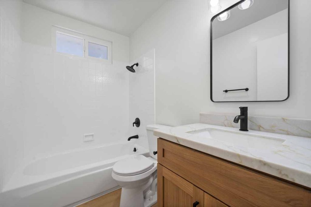 A bathroom with a light wood colored vanity with a white counter top, black fixtures, a tub/shower combo, and a toilet.