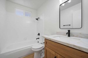 A bathroom with a light wood colored vanity with a white counter top, black fixtures, a tub/shower combo, and a toilet.