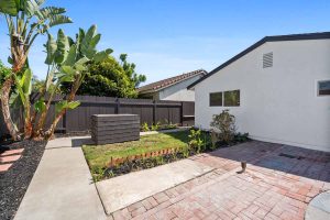 Backyard showing pavers, grassy area, white home and wooden fence.