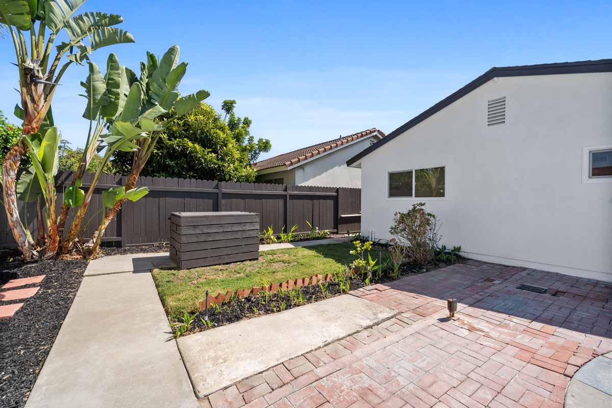 Backyard showing pavers, grassy area, white home and wooden fence.