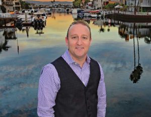 Danny Murphy wearing a blue and white gingham button up shirt with a black vest standing in front of the harbor.