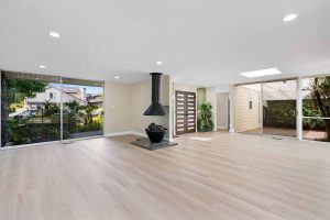 A large open space that is unfurnished showing a wood burning stove, mid-century modern double front doors with wooden slats, two sliding glass doors on either side of the room and new light wood flooring.