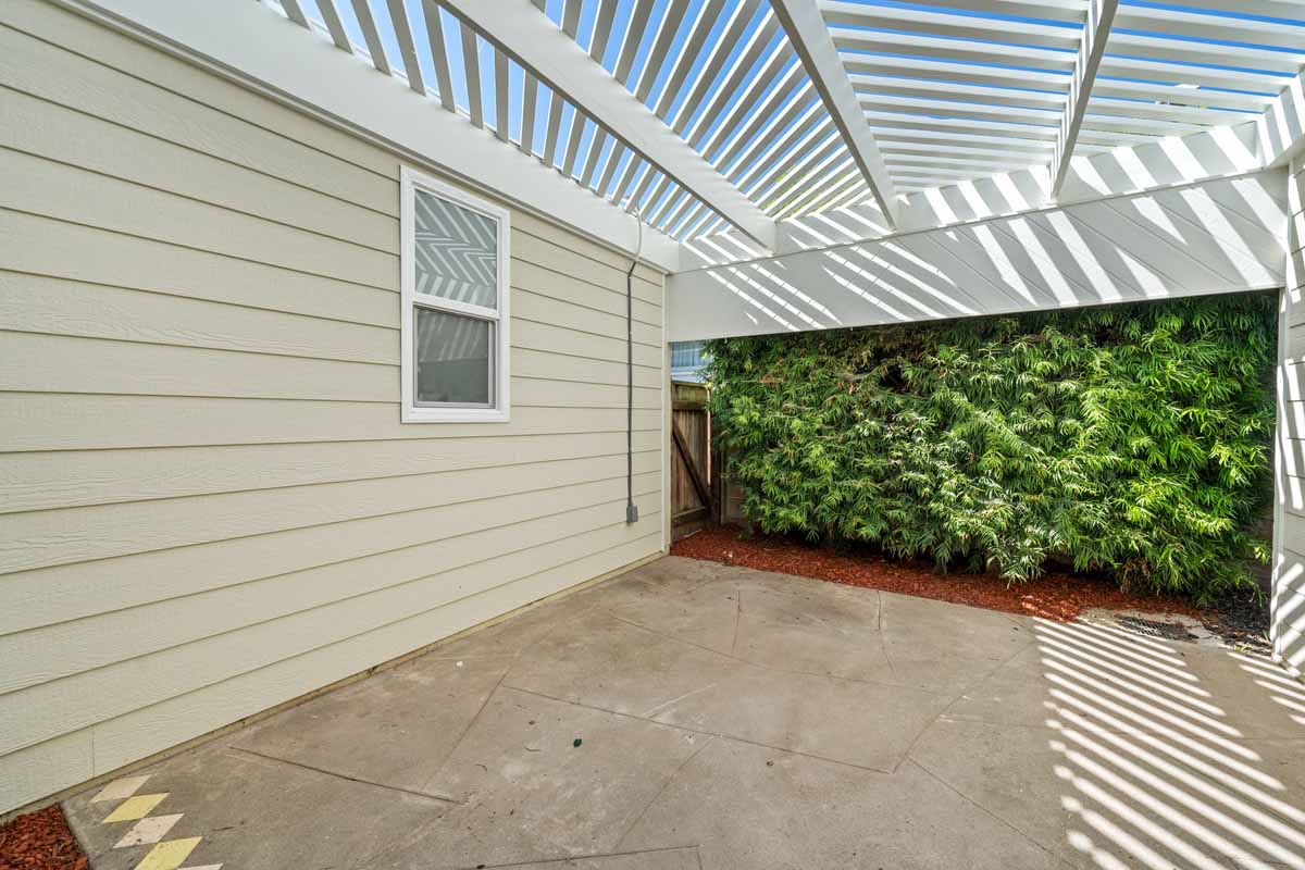 A white wood slatted covered patio with a wall to one side full of green foliage and a neutral painted wood siding with window ont he other side.
