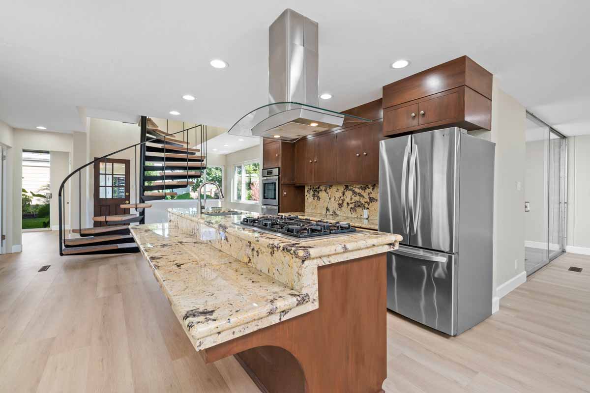 An updated kitchen showing a large, two-tiered island with beige colored granite, wood toned cabinets, and stainless steel appliances.