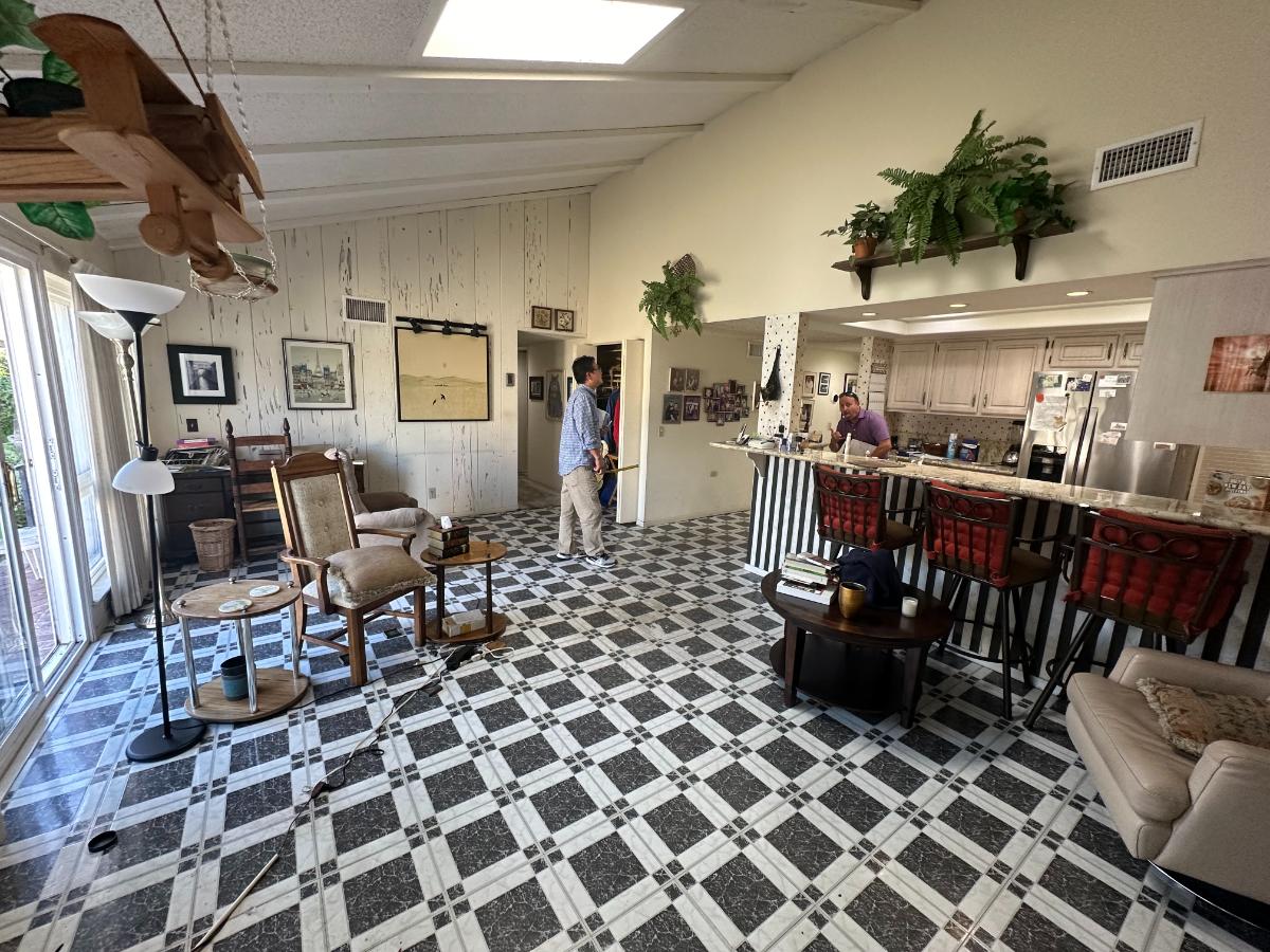 Before photo of a dining room with black and white flooring, a white wood accent wall, and a wallpapered kitchen. 