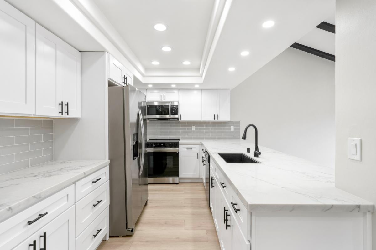 Bright new kitchen with wood tile flooring, bright white cabinets and walls, and white recessed lighting on the ceiling. 