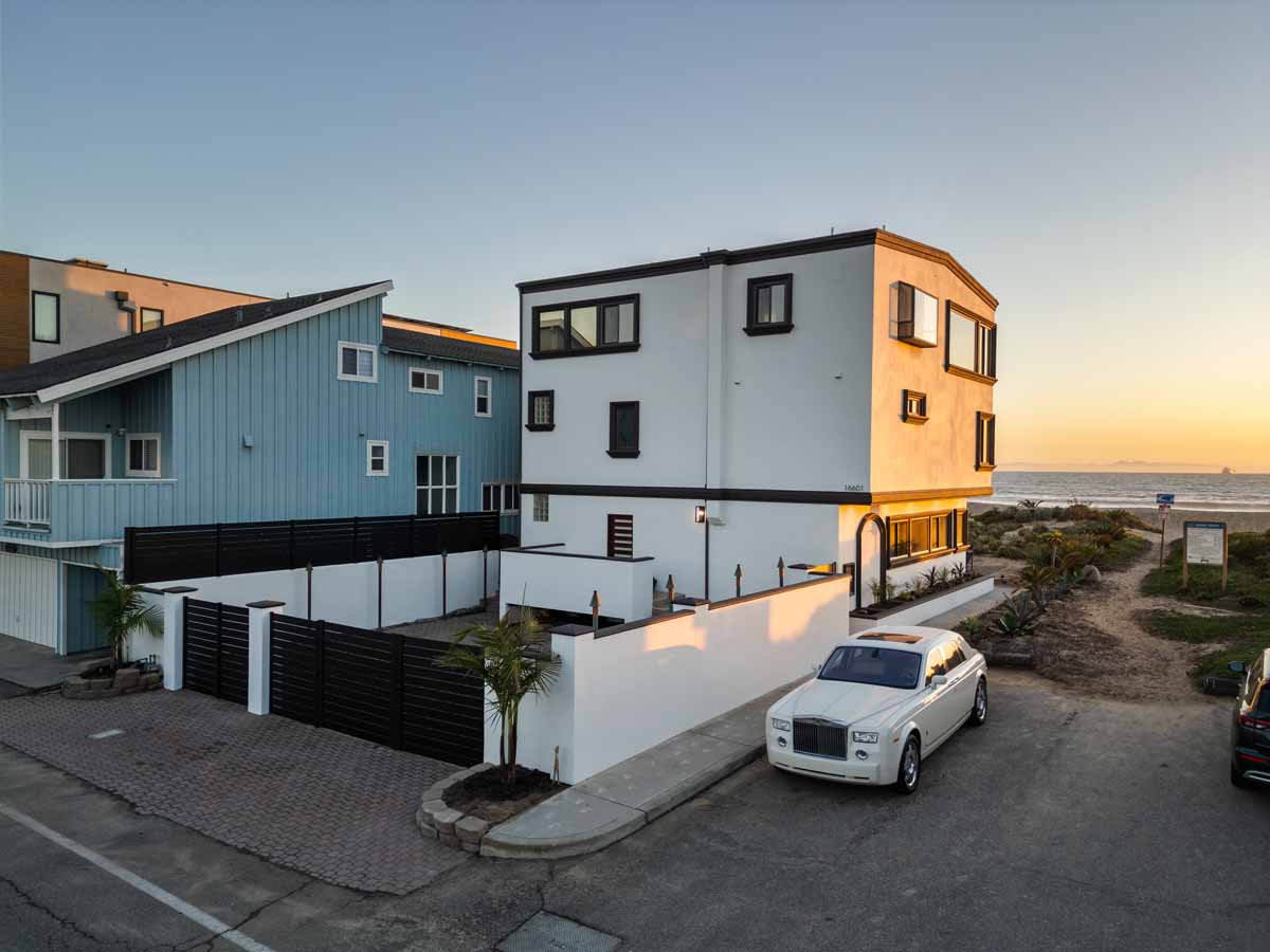 Exterior of white home showing a large gated parking area in the front and the beach and ocean in the background.
