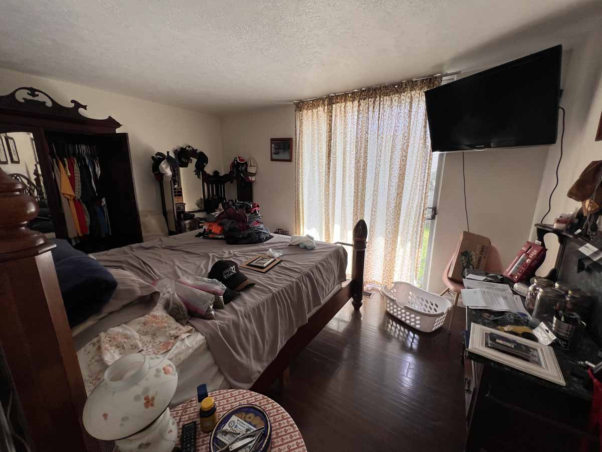 A cluttered bedroom with dark wood flooring, a bed, a lot of furniture and a mounted tv next to the covered sliding glass door.