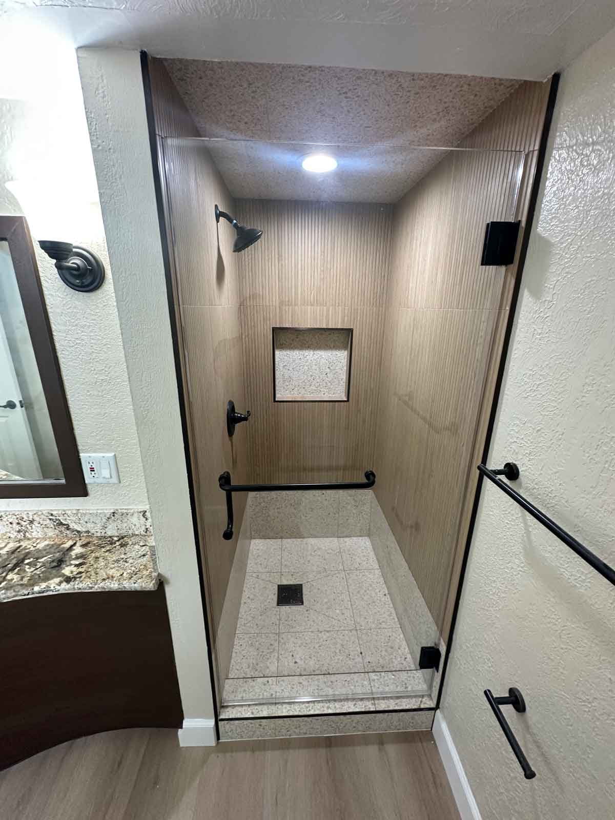 An updated bathroom showing a large, renovated walk-in shower with wood colored walls, new glass door and black fixtures.