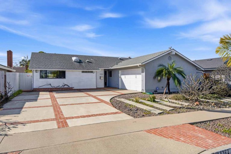 Exterior view of front of home showing a white and gray facade and brick inlaid into the concrete driveway.
