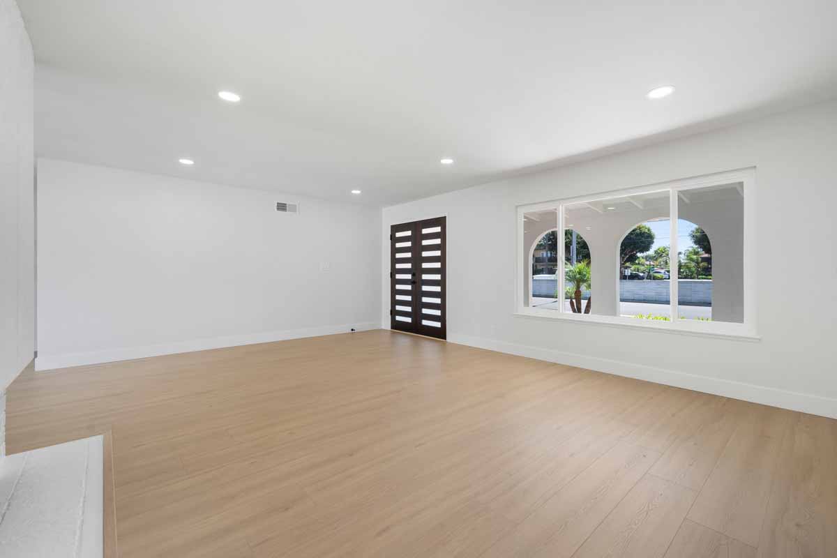 An updated living space with light colored wood flooring with white walls, new horizontal wood-slatted double front doors and a large window looking out at the front yard.