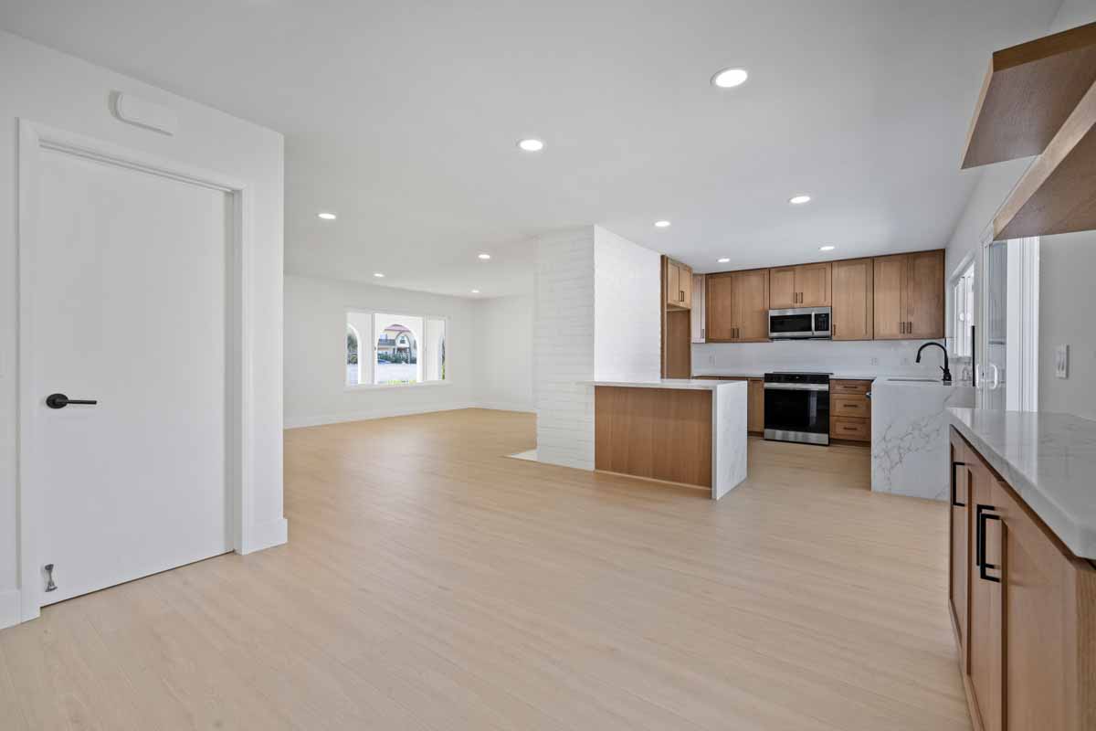 An updated dining space with light colored wood flooring, white walls, and a view into the updated kitchen with wood cabinetry and light stone counter tops.