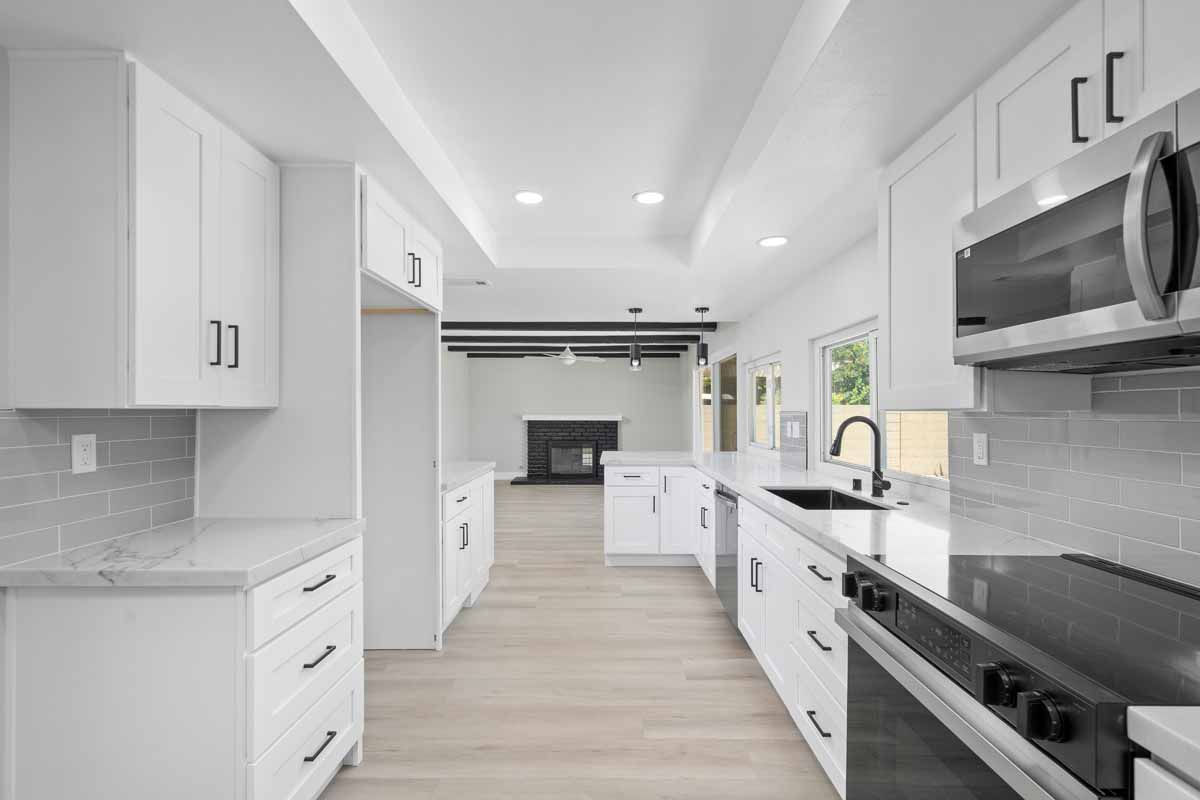 Updated kitchen with light wood colored flooring, neutral paint on walls, white cabinets and stone counters all looking towards the living room with a black fireplace.