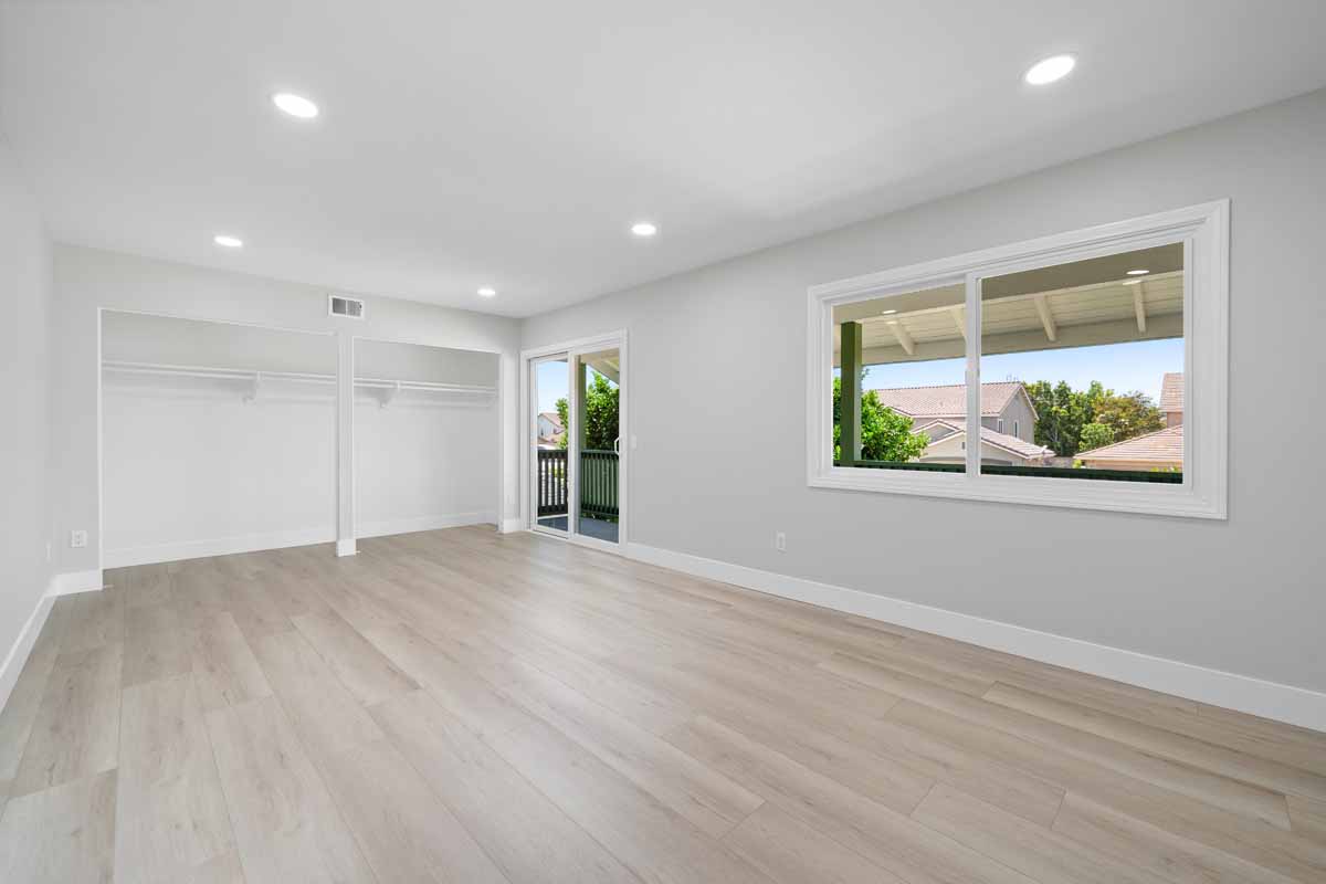 Large updated empty bedroom with light colored wood flooring, recessed lighting, neutral colored walls, a large window and a slider leading to second level balcony.