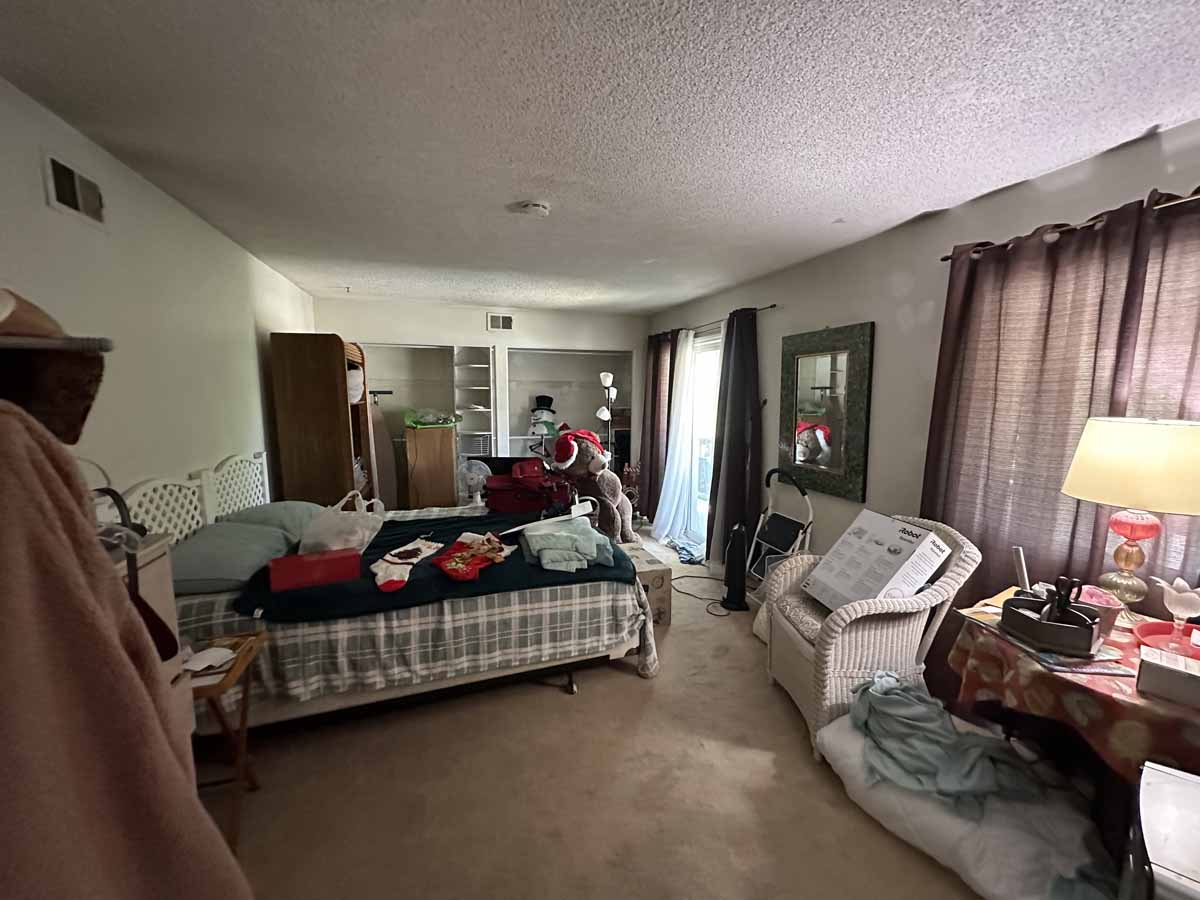Cluttered, outdated bedroom with brown carpet and dark curtains covering windows.