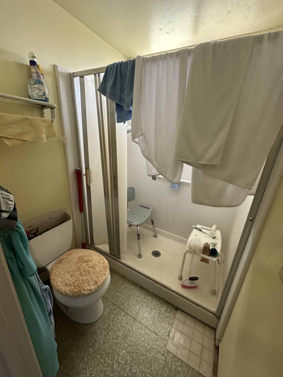 Outdated, cluttered bathroom with walk-in shower and a toilet.
