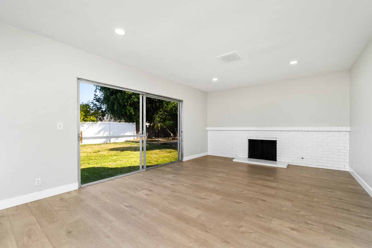 An empty living space with medium-toned wood flooring, neutral colored walls and a white fireplace, with a sliding glass door leading to the backyard.