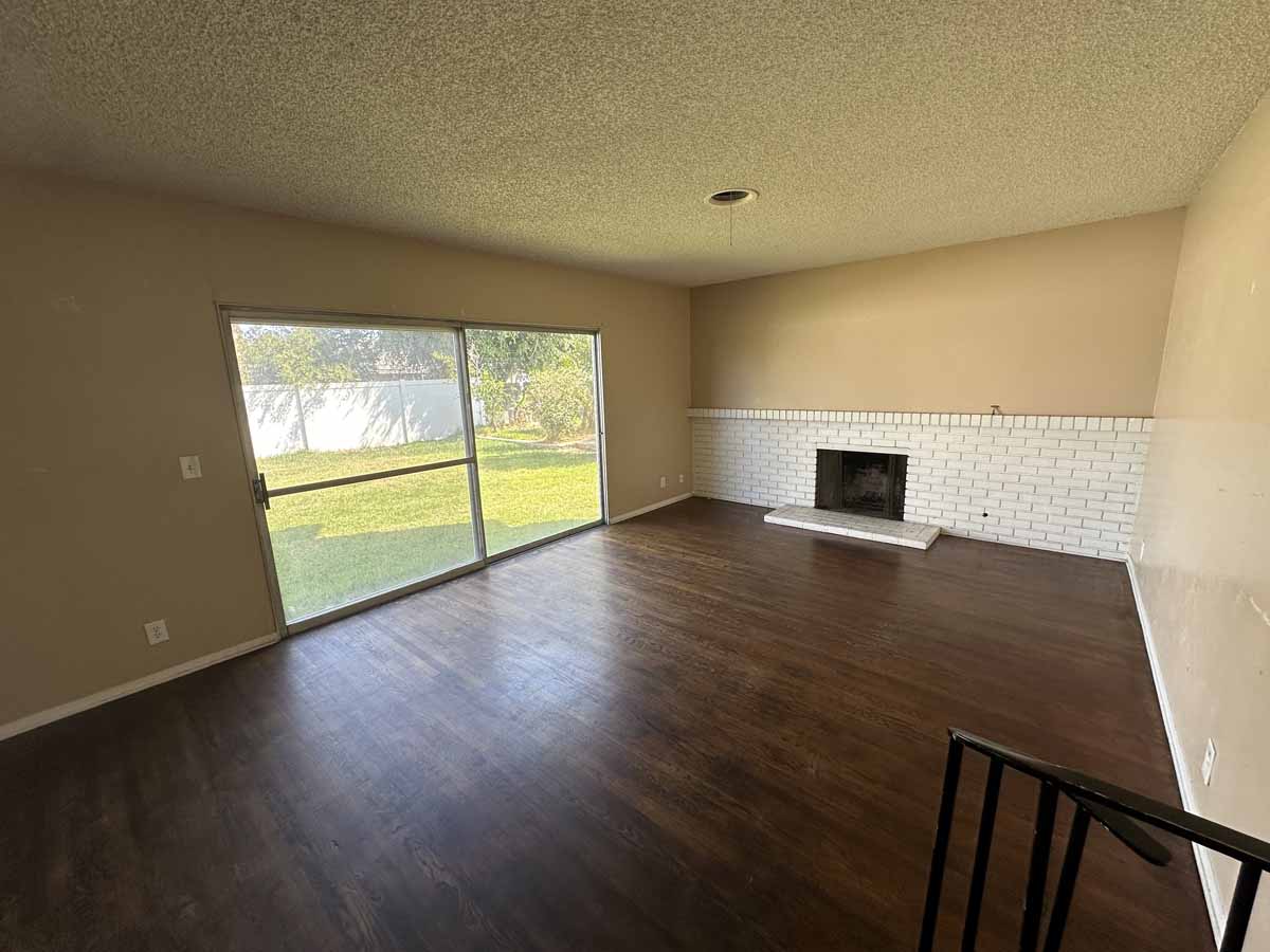 Darker wood planked flooring with a long white brick fireplace and yellow/cream toned wall paint and a sliding glass door leading to the backyard.