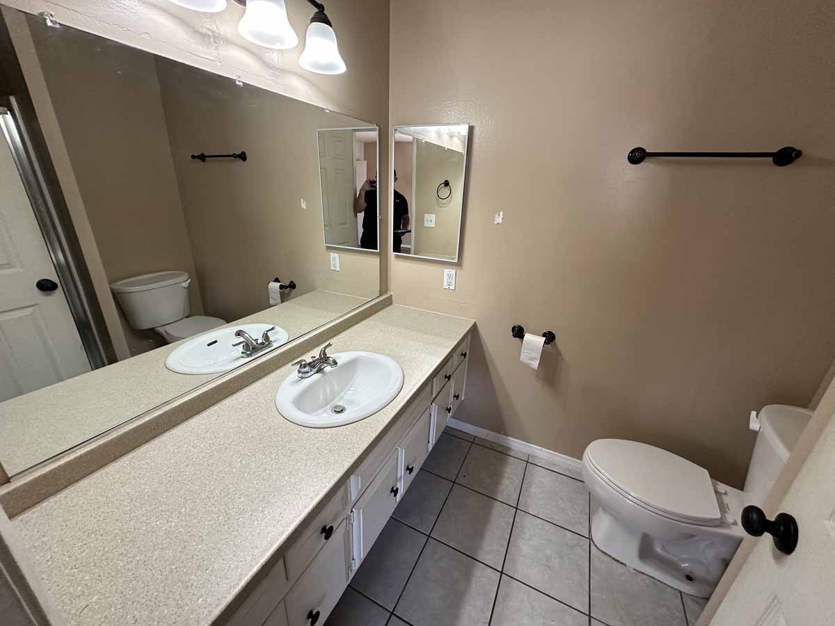 An outdated bathroom showing a long vanity with one sink, a large mirror above the vanity, and a toilet on the opposite side.