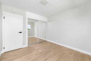 An empty bedroom with neutral colored walls, light-medium toned wood flooring and mirrored sliding closet doors.
