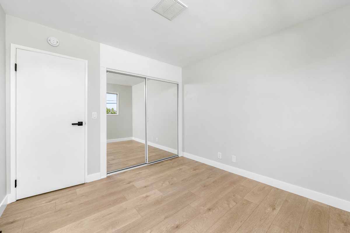 An empty bedroom with neutral colored walls, light-medium toned wood flooring and mirrored sliding closet doors.