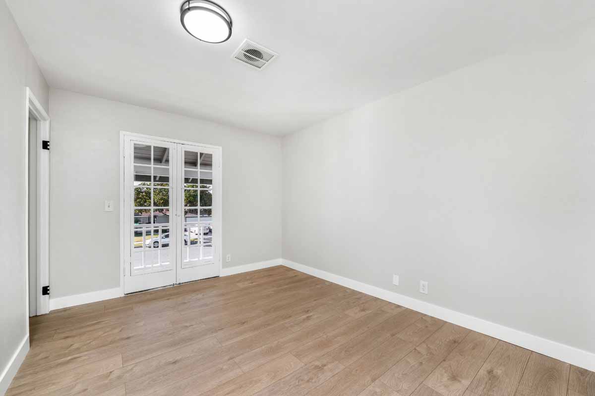 An empty bedroom with neutral colored walls, light-medium toned wood flooring and French doors leading outside.