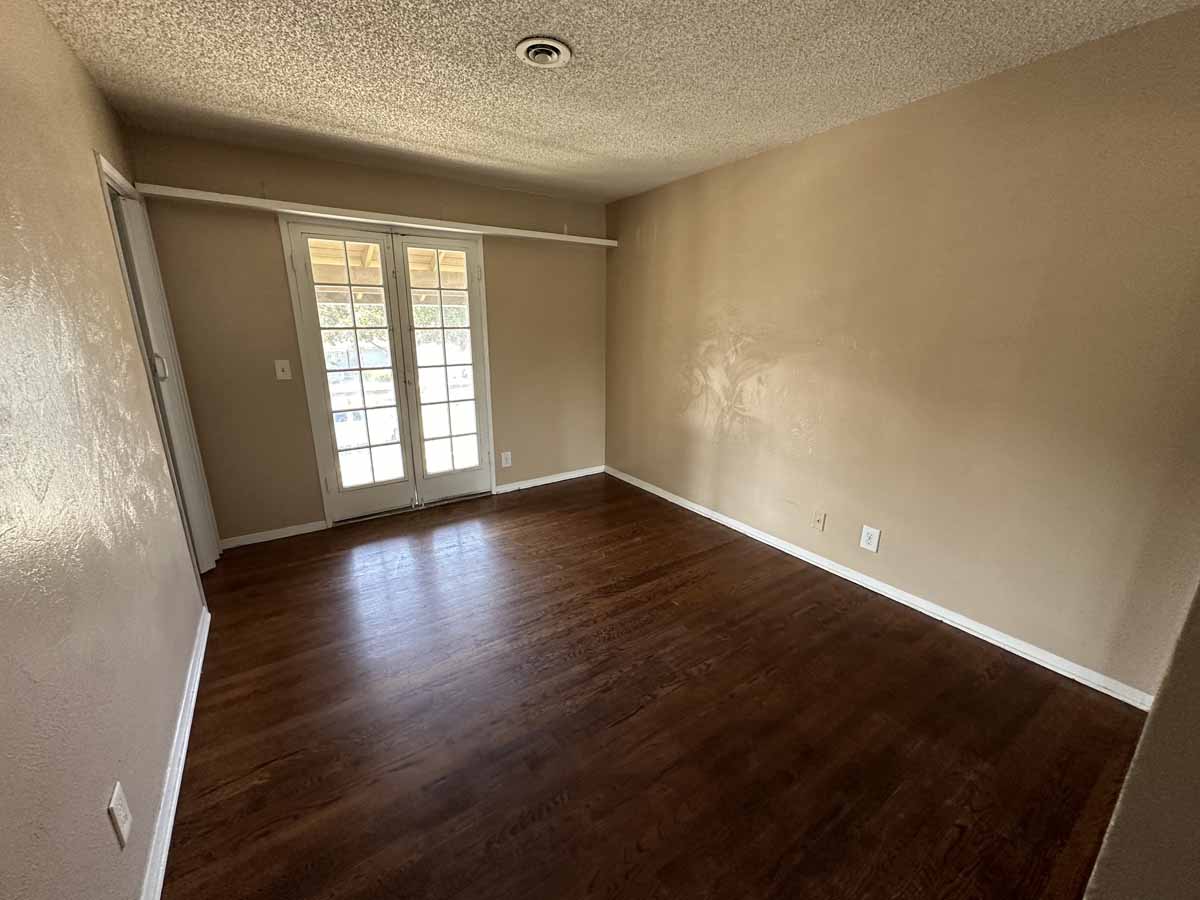 An empty bedroom with dark wood flooring, French doors leading to the backyard, and yellow/cream colored walls.