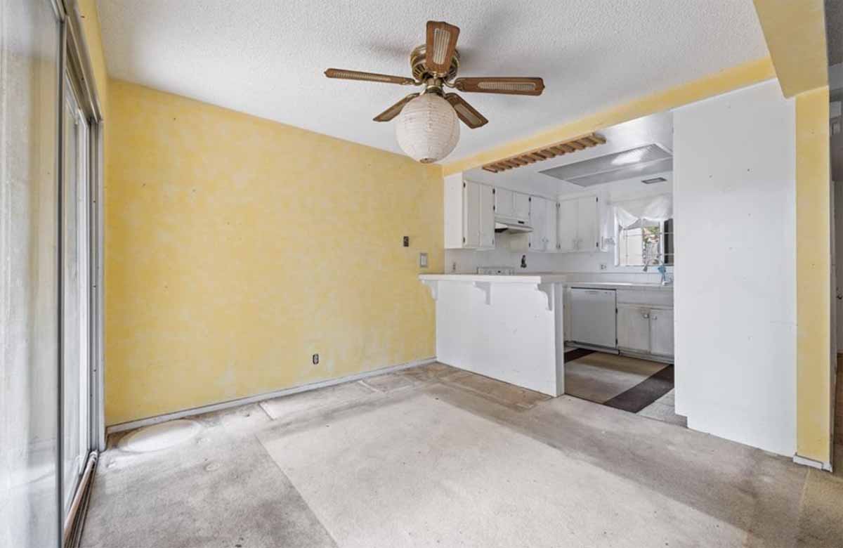 Outdated home with old beige carpet, yellow walls, ceiling fan and kitchen in the background.