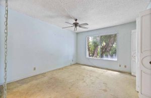 An empty bedroom with old beige carpet, a large window and a ceiling fan.
