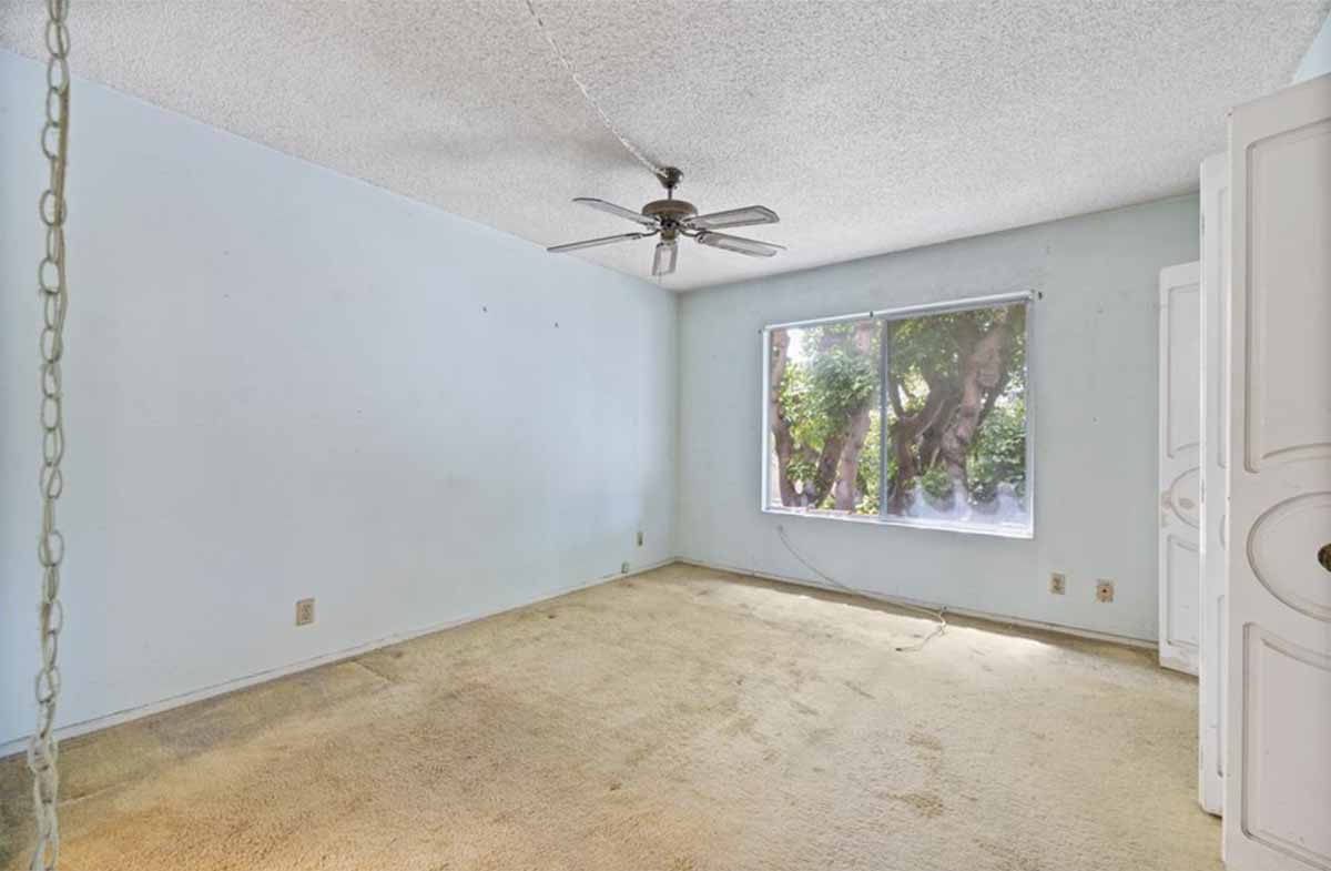 An empty bedroom with old beige carpet, a large window and a ceiling fan.