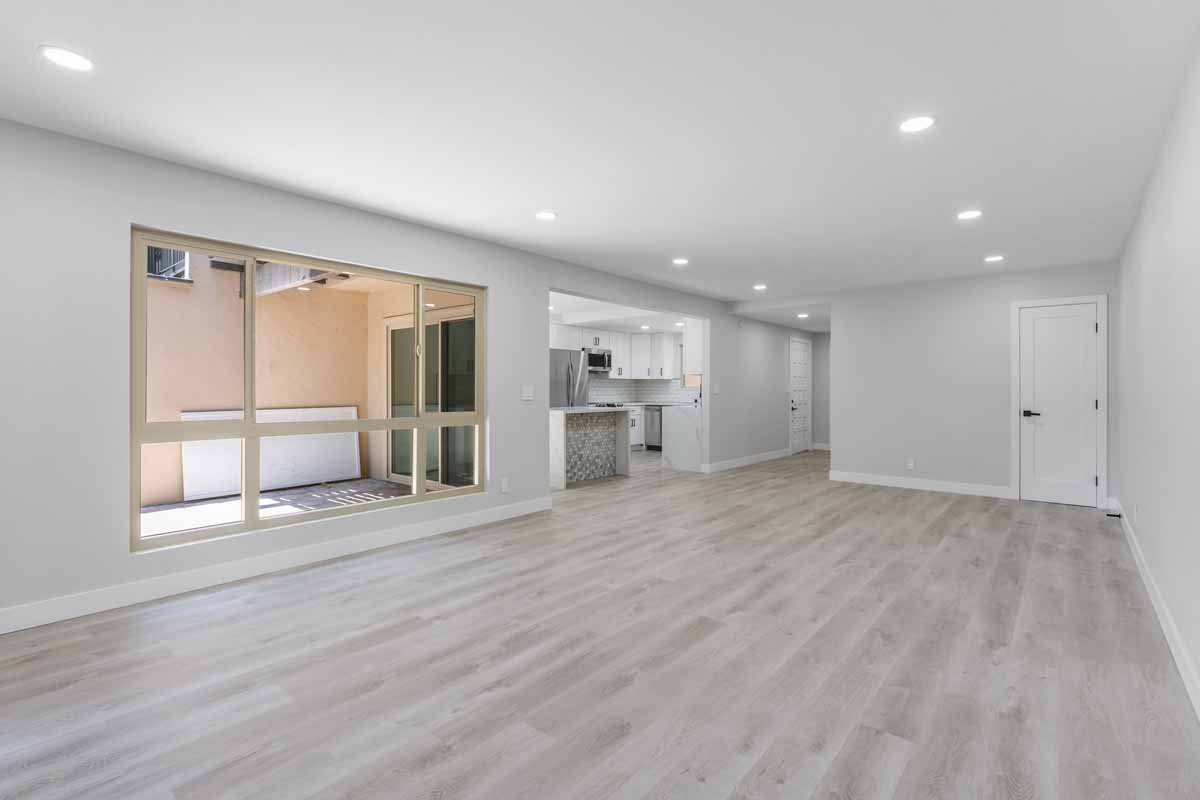 An updated living space with recessed lighting, neutral color walls and wood plank floors, a sliding glass door and the kitchen in the background.