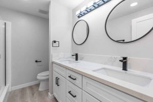 An updated bathroom with a two sink white vanity with white stone counter tops, two circle mirrors and a toilet in the background.