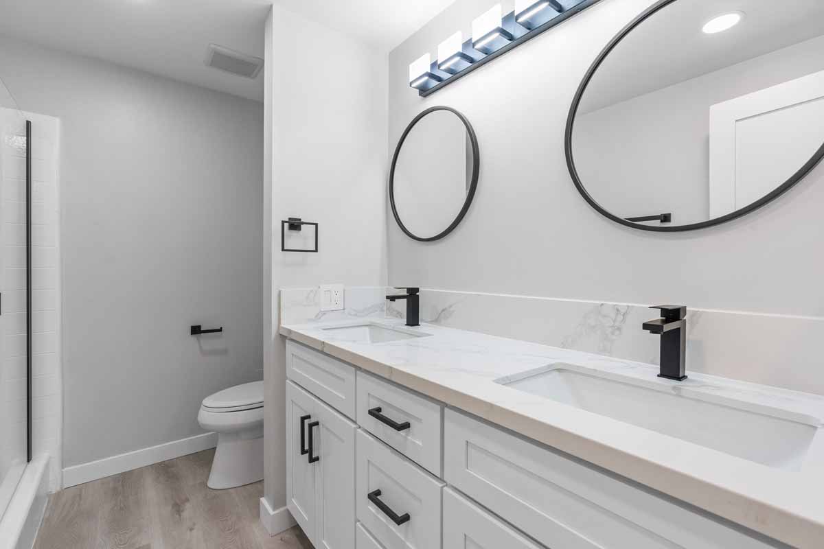 An updated bathroom with a two sink white vanity with white stone counter tops, two circle mirrors and a toilet in the background.