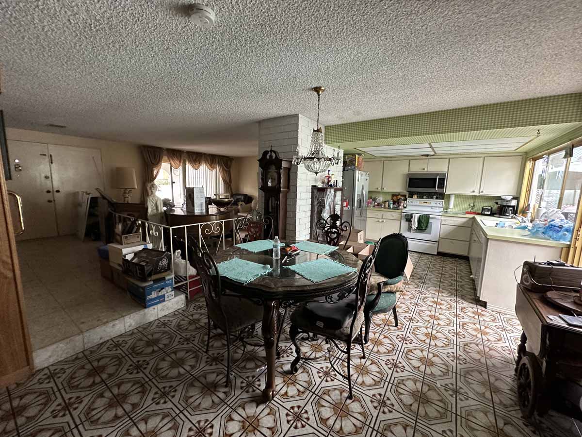 An outdated dining space that is open to both the kitchen and living room. The floors are a brown and cream, 70s style and there is an old dining table and chairs.