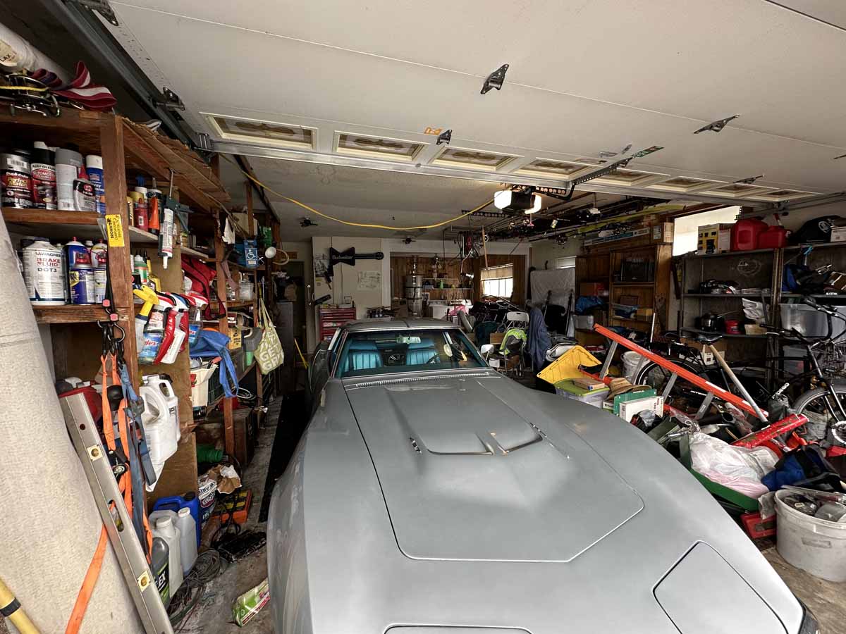 A cluttered garage showing an old car surrounded by shelves of paint, cleaners, etc.