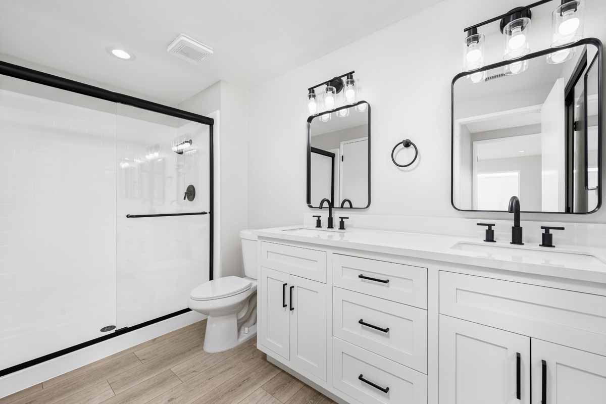 An updated bathroom with a two sink white vanity with white stone counter tops, two mirrors and a toilet and shower in the background.