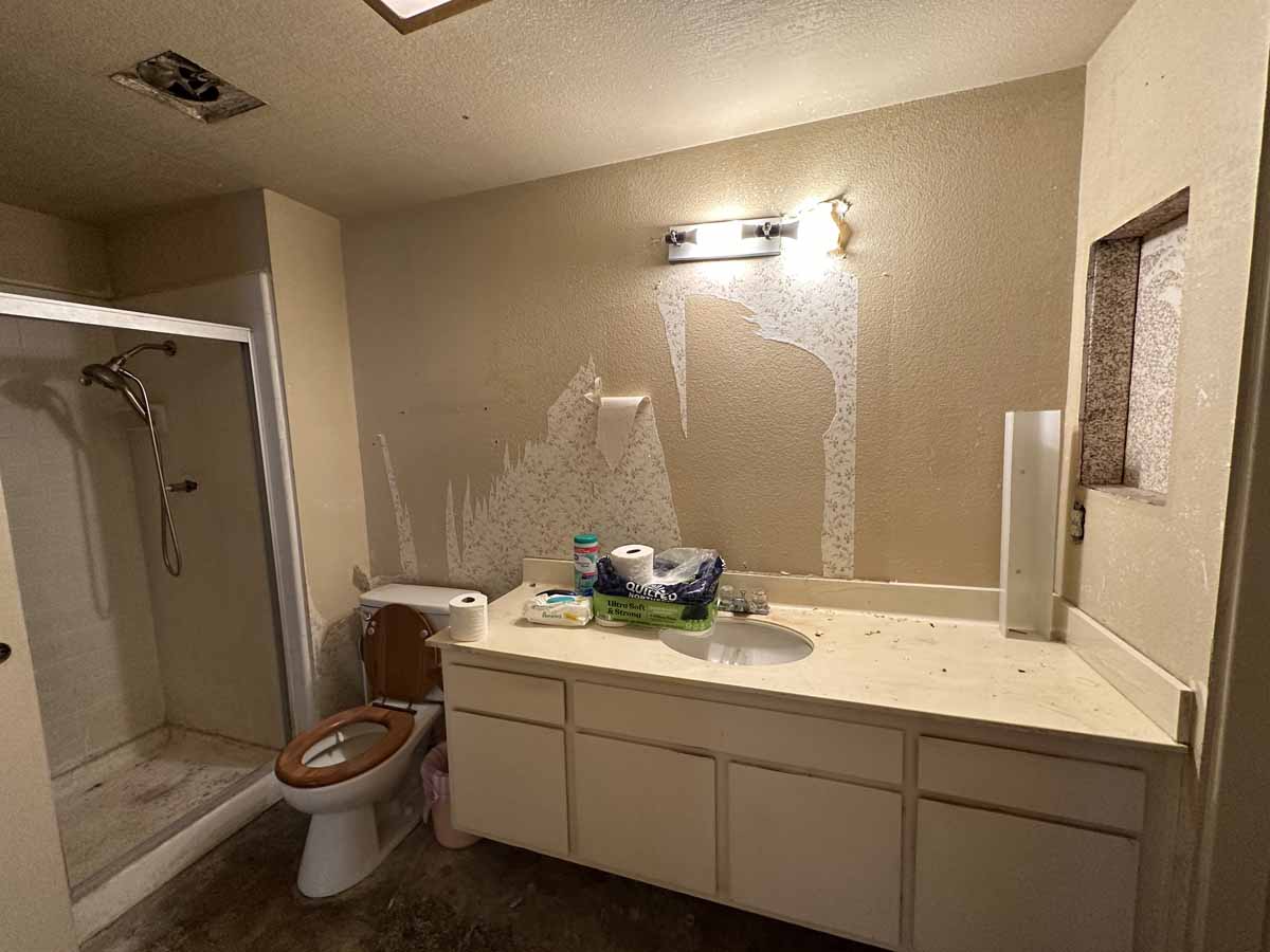 Outdated bathroom with step-in shower, toilet with wooden seat and single sink vanity.