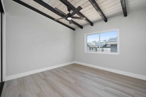 An empty bedroom with light wood flooring, light neutral wall paint, wood beams on the ceiling and a window.