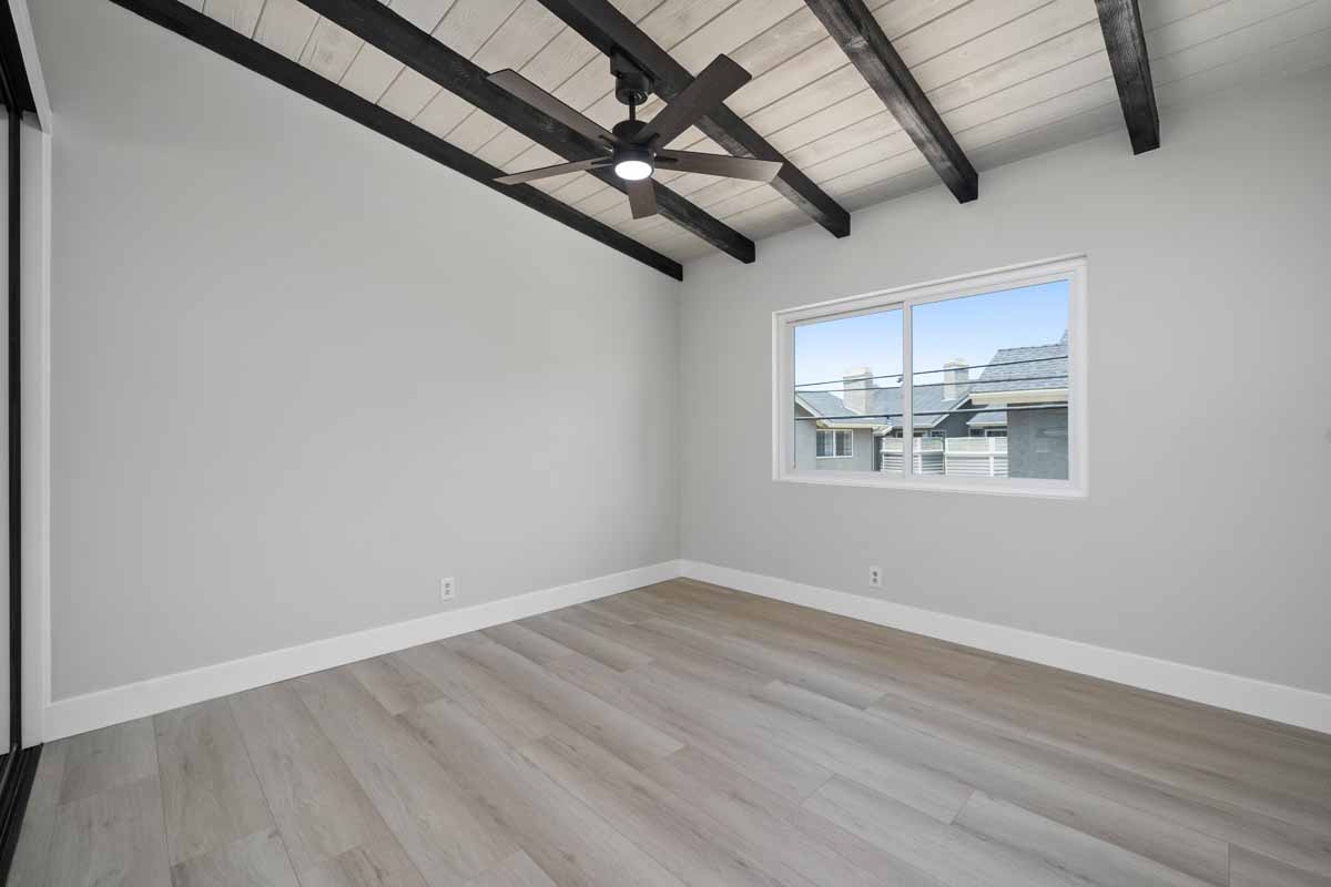 An empty bedroom with light wood flooring, light neutral wall paint, wood beams on the ceiling and a window.