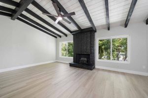 An empty living space with light wood flooring and a black fireplace on the back wall flanked by two large windows. The ceiling has light wood planks and dark wood beams.