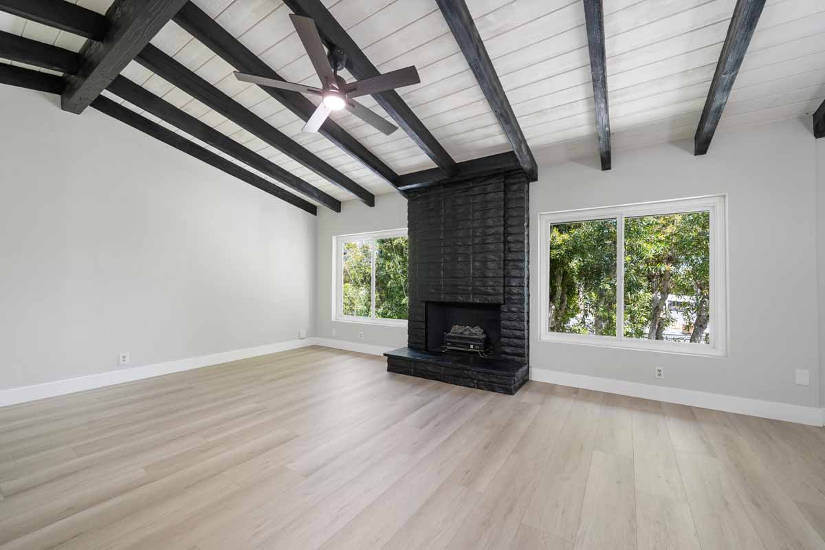 An empty living space with light wood flooring and a black fireplace on the back wall flanked by two large windows. The ceiling has light wood planks and dark wood beams.