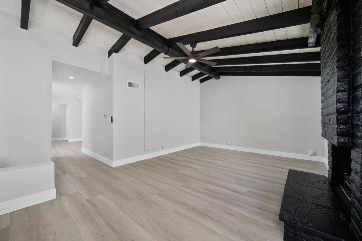 An empty living space with light wood flooring and a black fireplace off to the right with dark wood beams on the ceiling.